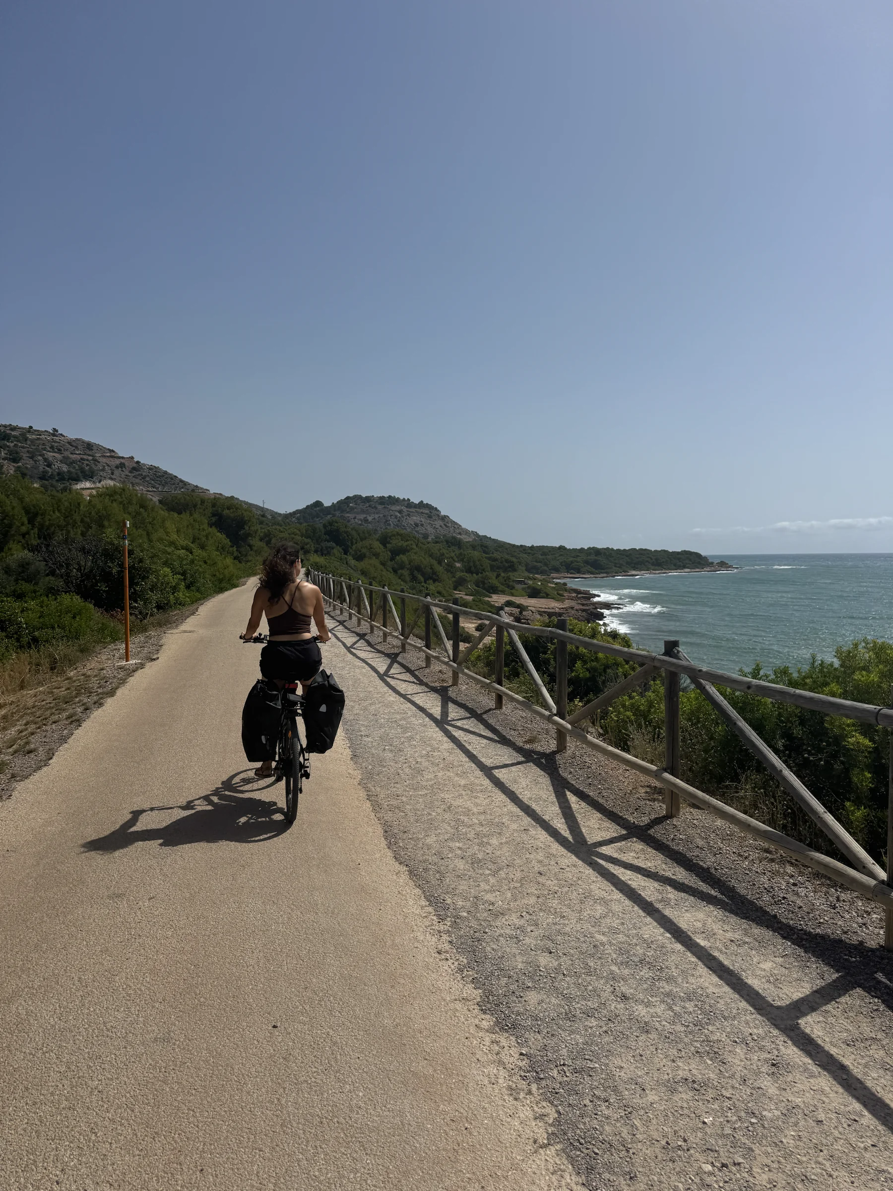 A cyclist riding a loaded bicycle along a quiet Mediterranean coastal path.