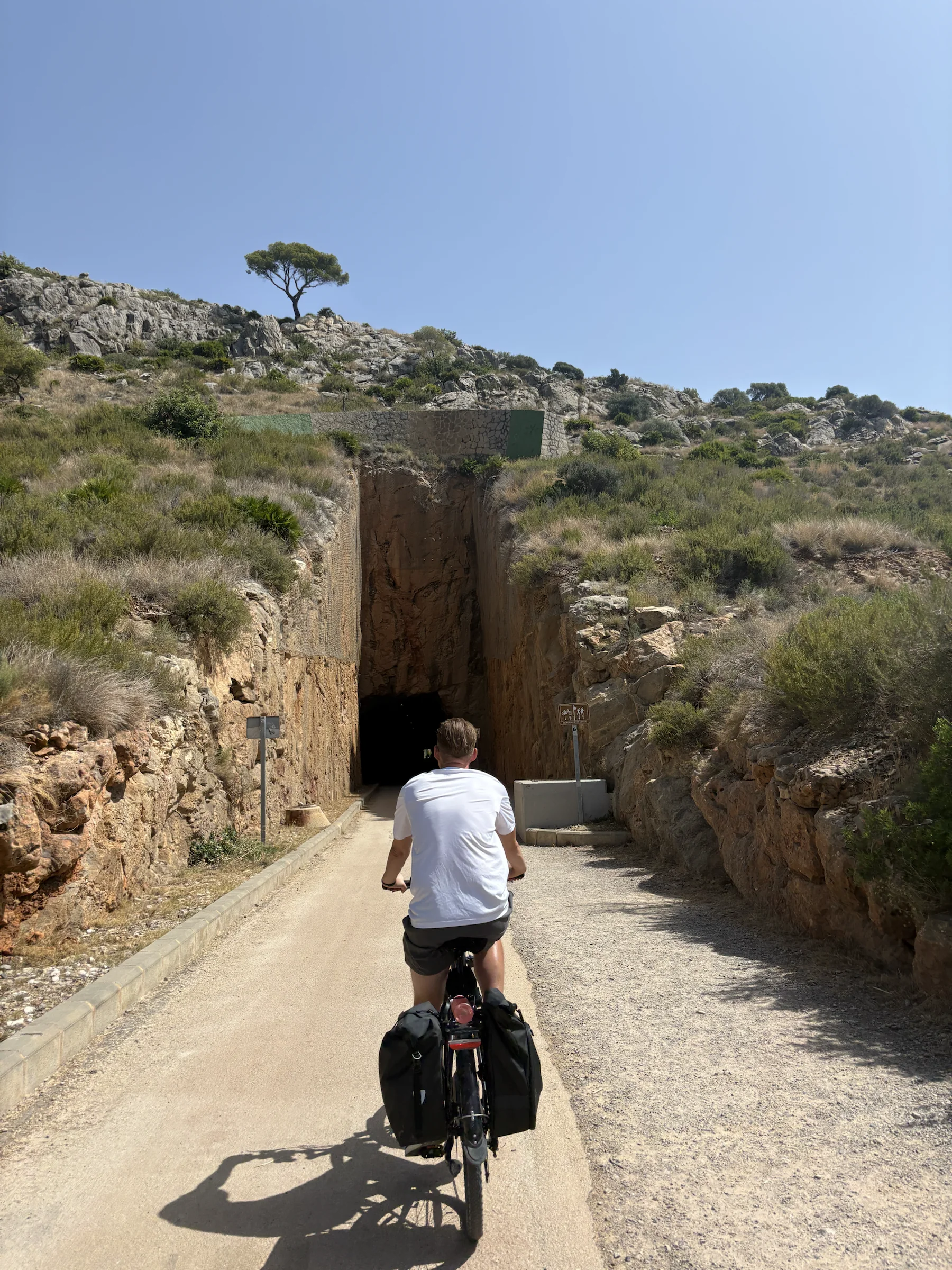 A rider on a loaded bicycle heading toward a tunnel cut through the rocky coastal hillside.
