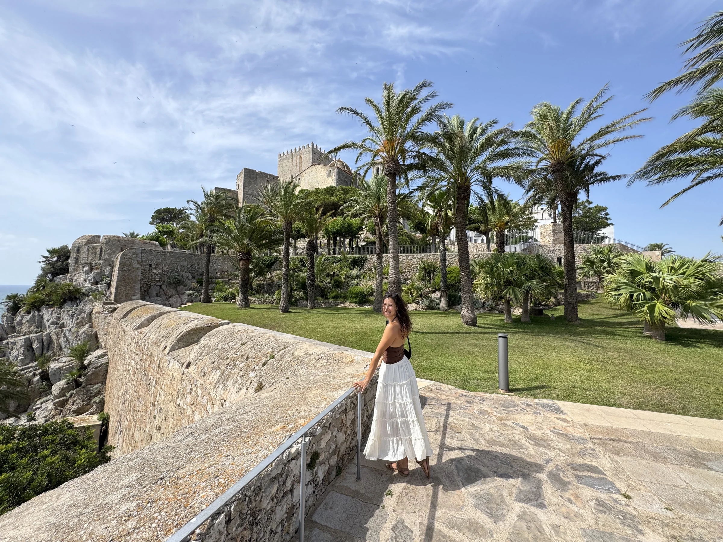 Traveller on a palm-lined path beside Peñíscola castle and the sea.